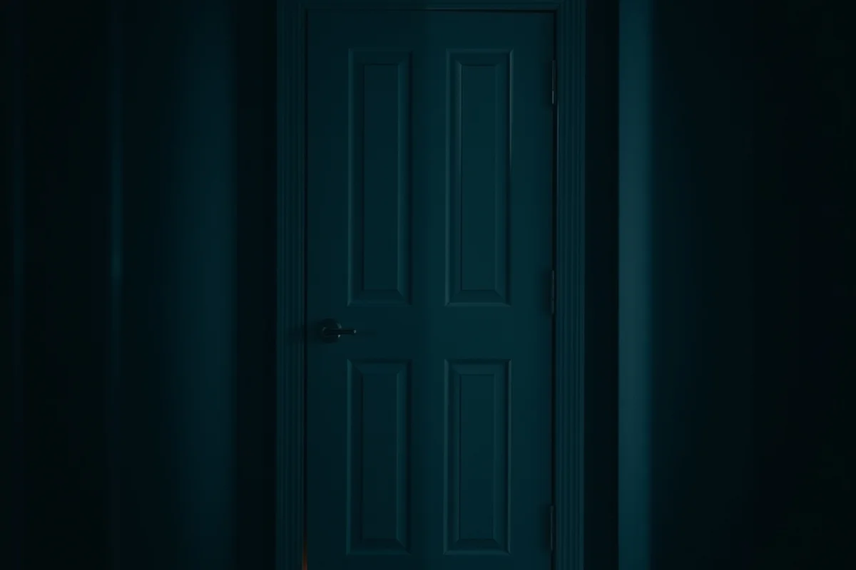 una porta chiusa in un corridoio in ombra, un filo di luce calda che filtra dal basso